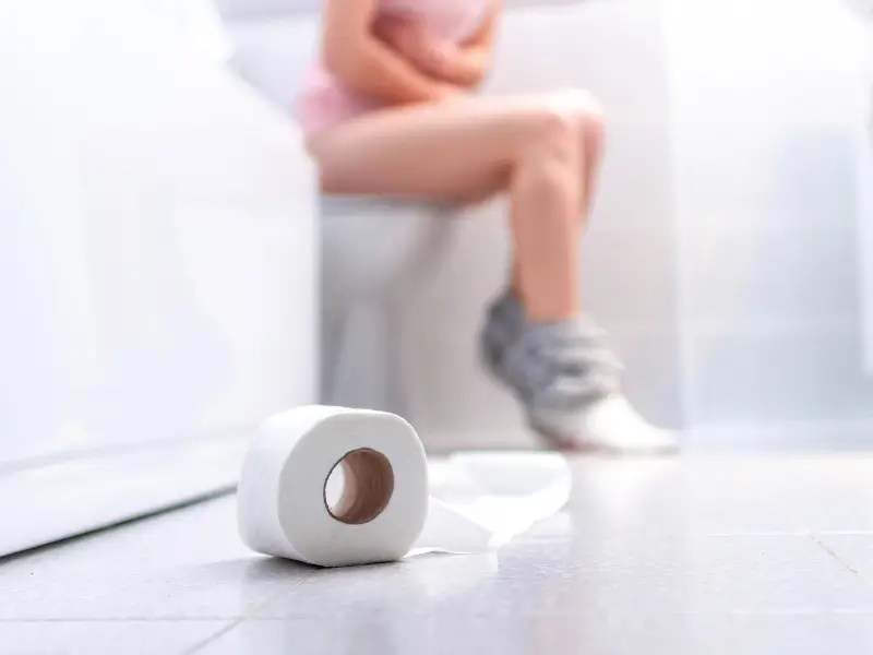 Person experiencing bowel discomfort while sitting on the toilet with a toilet paper roll on the floor.